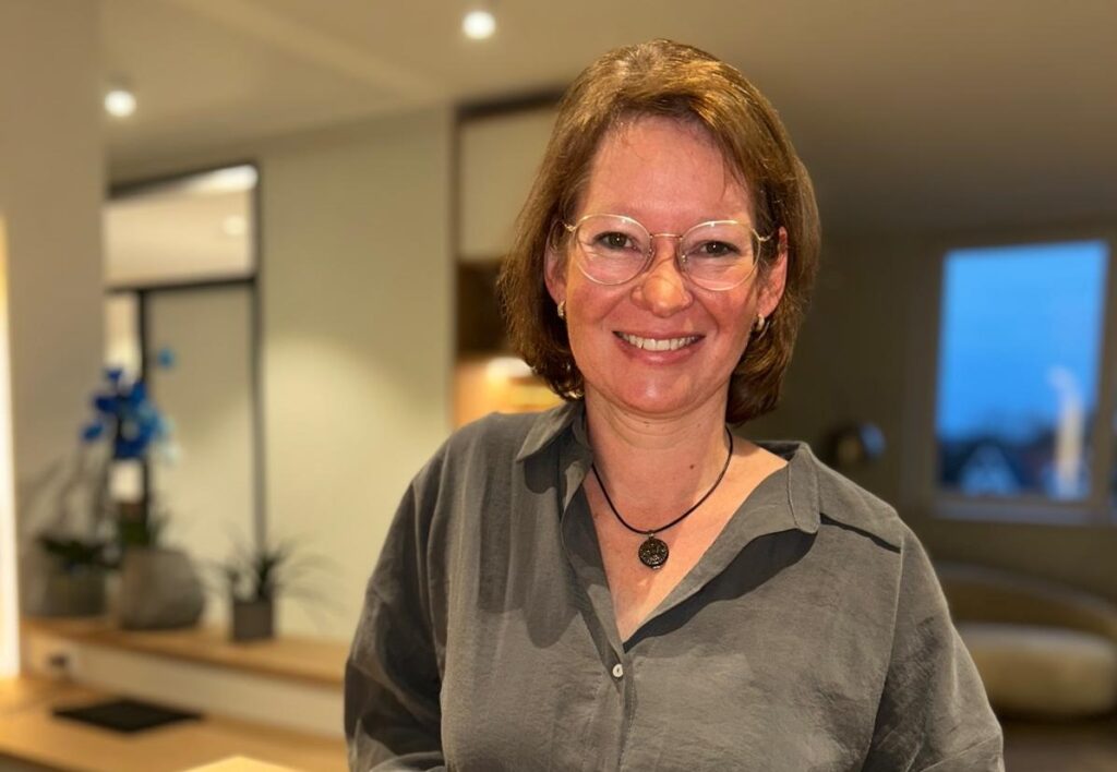 A woman with short brown hair and glasses smiles warmly indoors. She is wearing a grey shirt and a necklace, with a softly lit modern room in the background.
